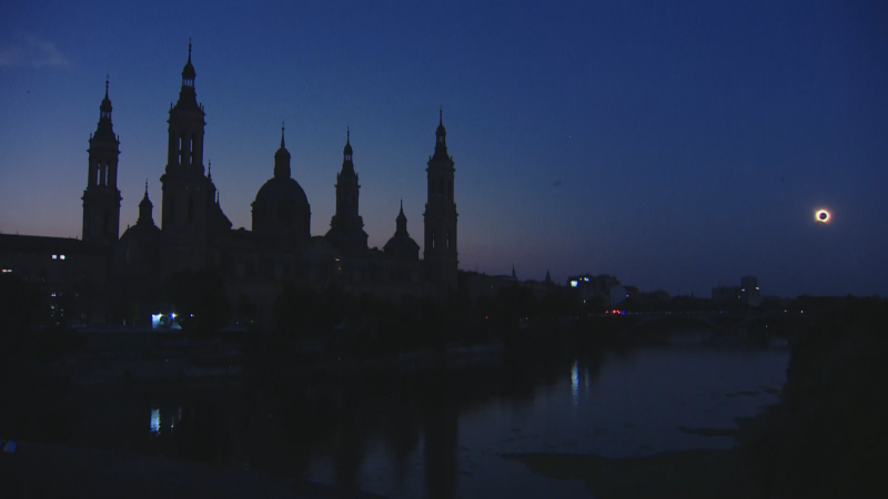 Vista nocturna de Zaragoza con la Basílica del Pilar y el río Ebro. Un eclipse solar anular ilumina el cielo oscuro, creando un efecto de anillo de diamante.