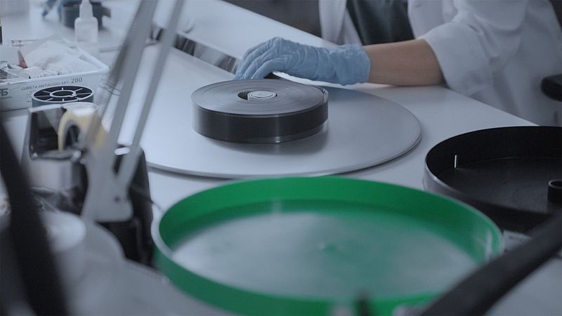 Manos enguantadas de azul trabajan con un carrete de película cinematográfica en un laboratorio, rodeadas de equipo de restauración y suministros.