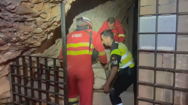 Miembros de la UME y personal "PM" trabajan juntos moviendo un objeto en una cueva o estructura rocosa. Se aprecian uniformes rojos con franjas amarillas y un chaleco reflectante.