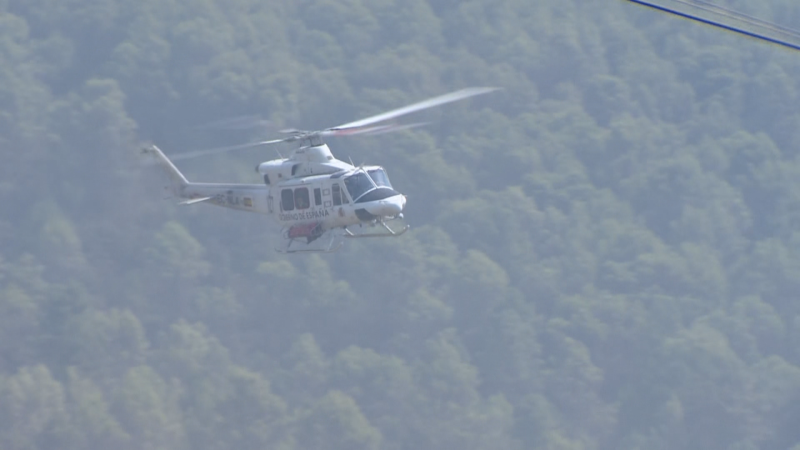 Helicóptero de emergencias blanco y rojo en vuelo sobre un paisaje montañoso y boscoso. "EMERGENCIAS" visible en el lateral.