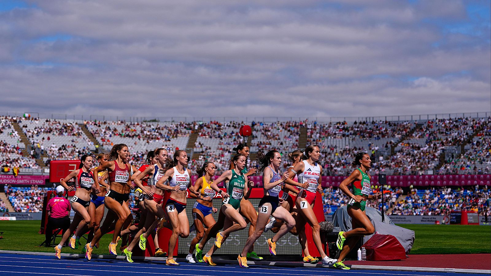 Carrera de atletismo femenino con atletas en acción en pista azul, luciendo uniformes con dorsales y patrocinadores como NISSAN. Se aprecian inscripciones de países como IRELAND y FRANCE.