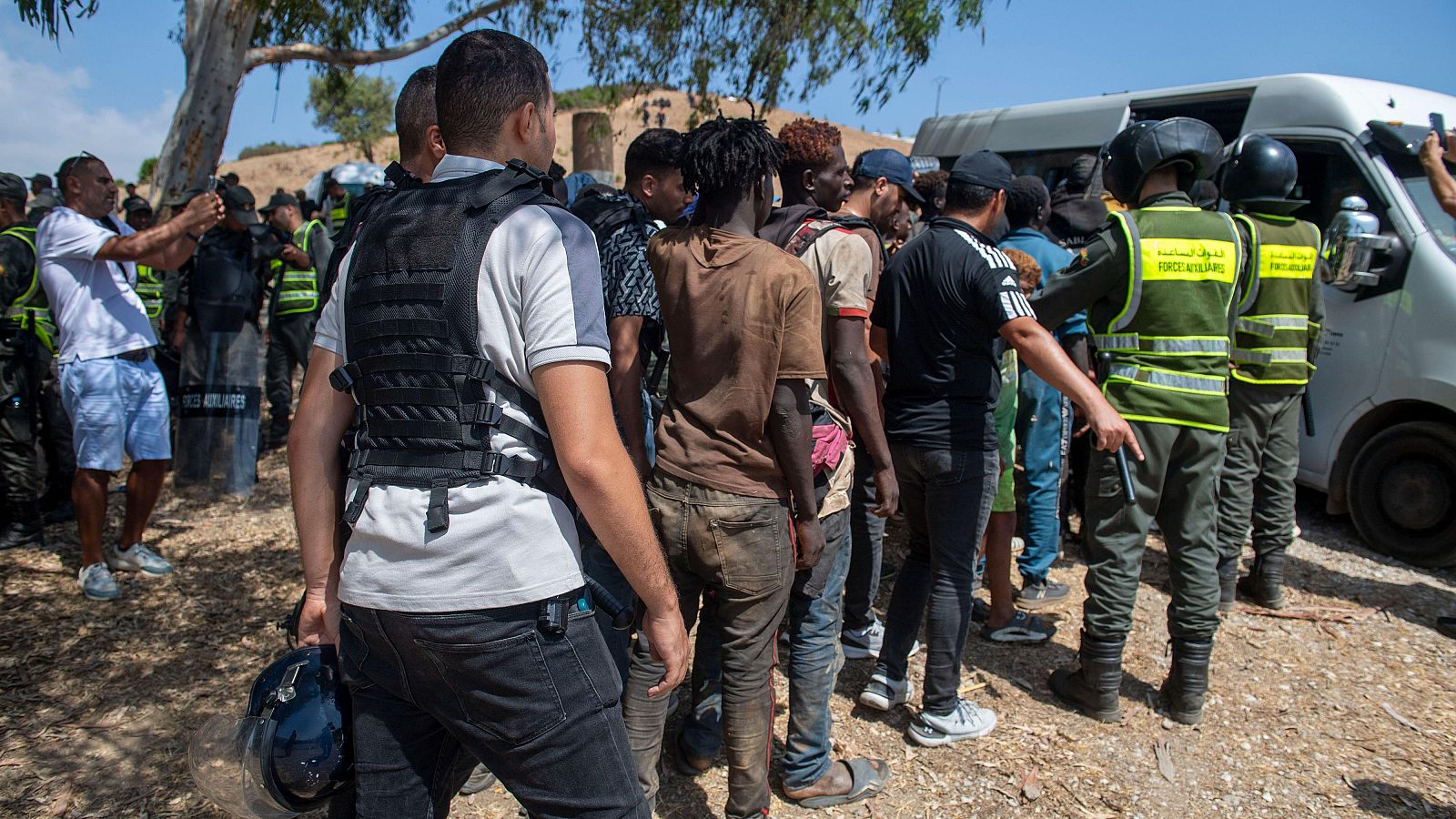 Fuerzas de seguridad marroquíes dirigen a hombres de ascendencia africana en un día soleado. Se aprecian uniformes con texto en árabe "القوات المساعدة" y "الدرك الملكي".