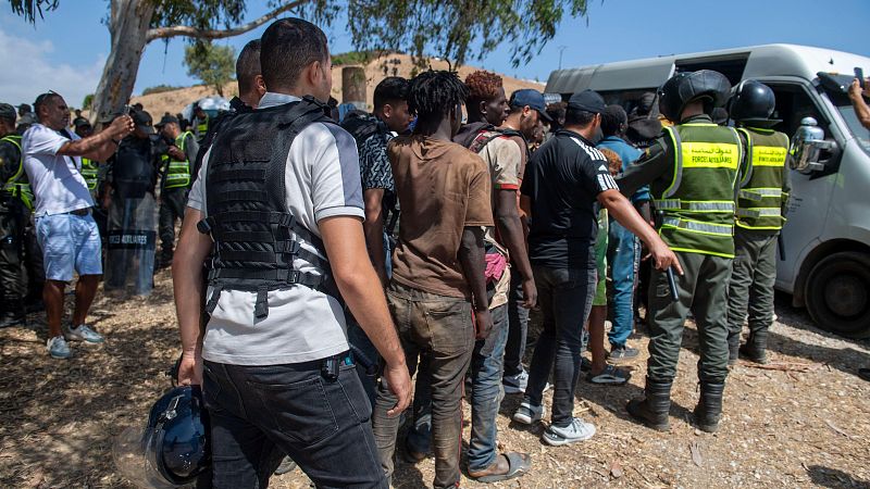 Fuerzas de seguridad marroquíes dirigen a hombres de ascendencia africana en un día soleado. Se aprecian uniformes con texto en árabe "القوات المساعدة" y "الدرك الملكي".