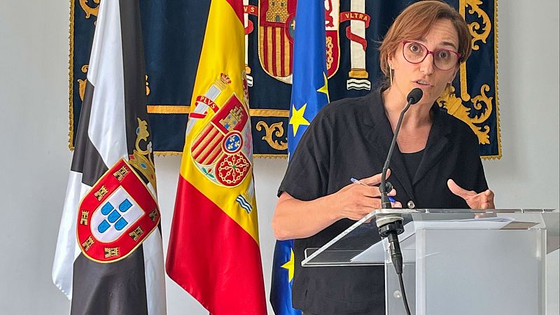 Yolanda Díaz habla en un atril con micrófono, flanqueada por las banderas de Ceuta, España y la UE. Lleva gafas rojas y blusa negra.