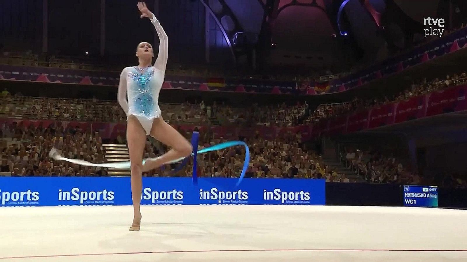 Alina Harnasko ejecuta un ejercicio de gimnasia rítmica con cinta turquesa en un pabellón deportivo ante un público. Se observa el logo 'rtve play' y 'inSports'.