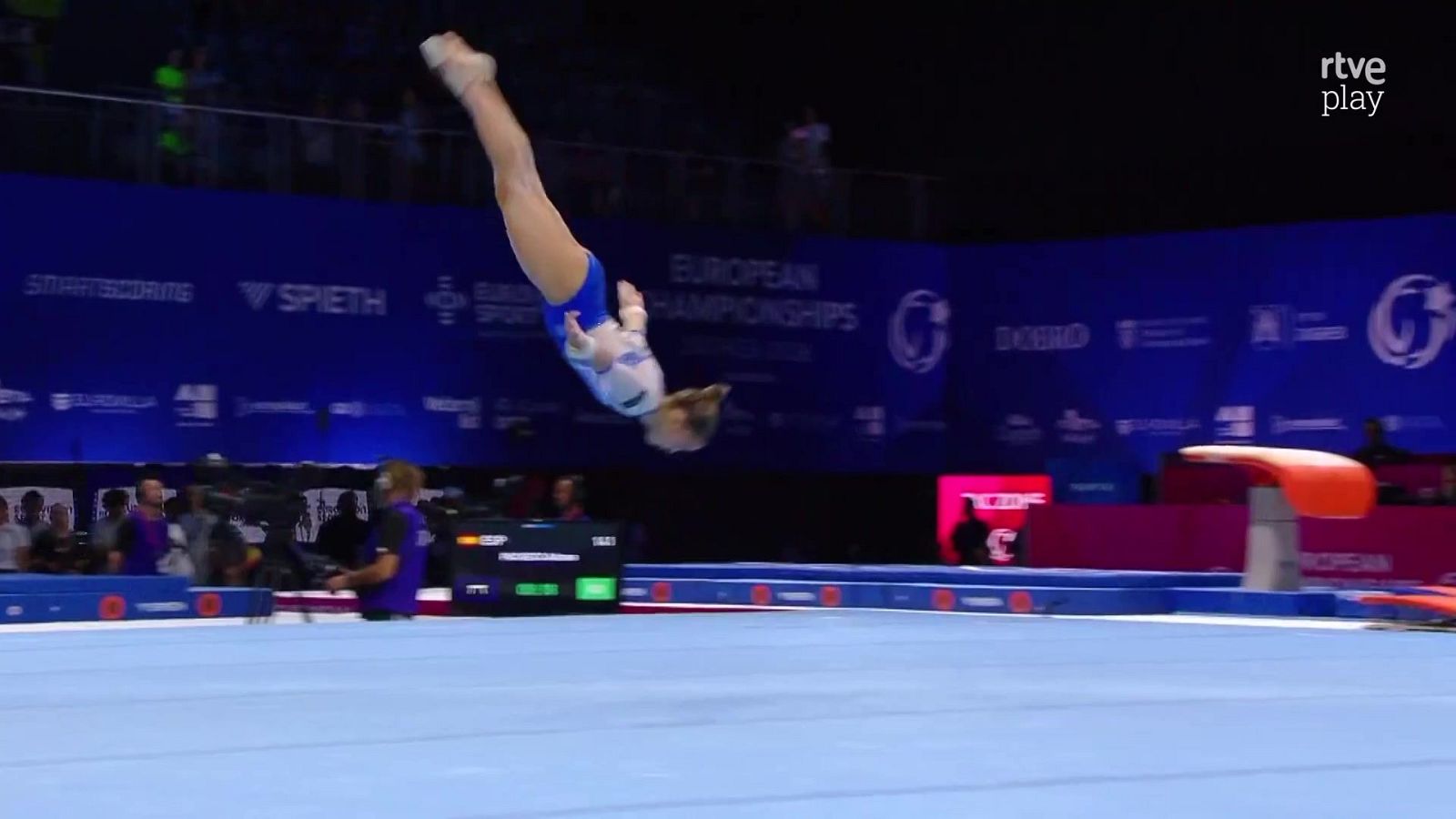 Gimnasta en pleno vuelo durante el Campeonato Europeo, con maillot blanco y azul. Se aprecian "EUROPEAN CHAMPIONSHIPS", "SMARTSCORING" y "rtve play".