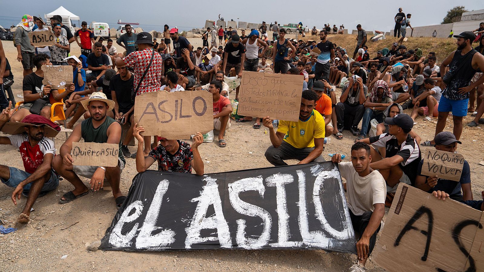 Hombres protestan en terreno árido exigiendo asilo, con carteles que denuncian la situación en Melilla y piden "No mas muertes. Asilo ya."