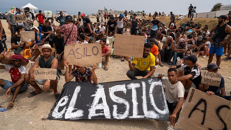 Hombres protestan en terreno árido exigiendo asilo, con carteles que denuncian la situación en Melilla y piden "No mas muertes. Asilo ya."