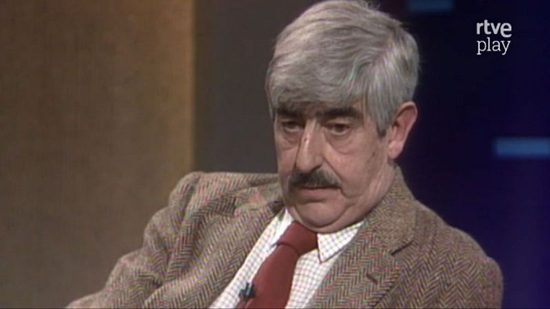 Retrato de Juan Benet, escritor, con traje de tweed y corbata roja, en un estudio de RTVE. Logo de "rtve play" visible.