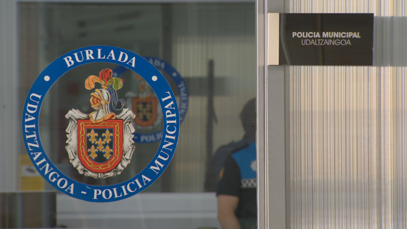 Entrada a la Policía Municipal de Burlada con puerta de cristal, logo circular azul y cartel "POLICIA MUNICIPAL - UDALTZAINGOA".