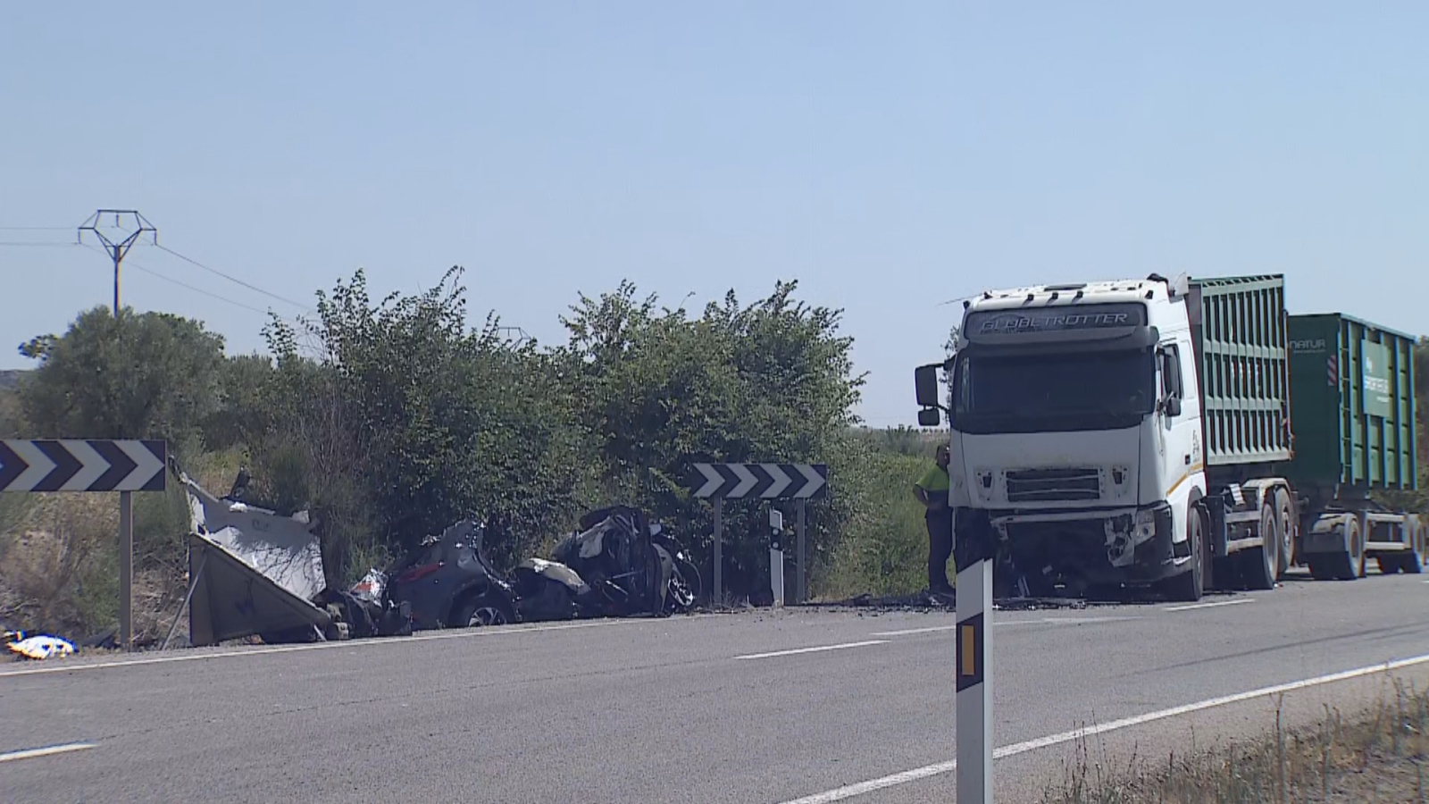 Accidente grave en carretera: camión 'CAMION TROTTER' y coche destrozado. Señal de curva peligrosa y personal de emergencia presentes. Entorno rural con vegetación y torre eléctrica al fondo.