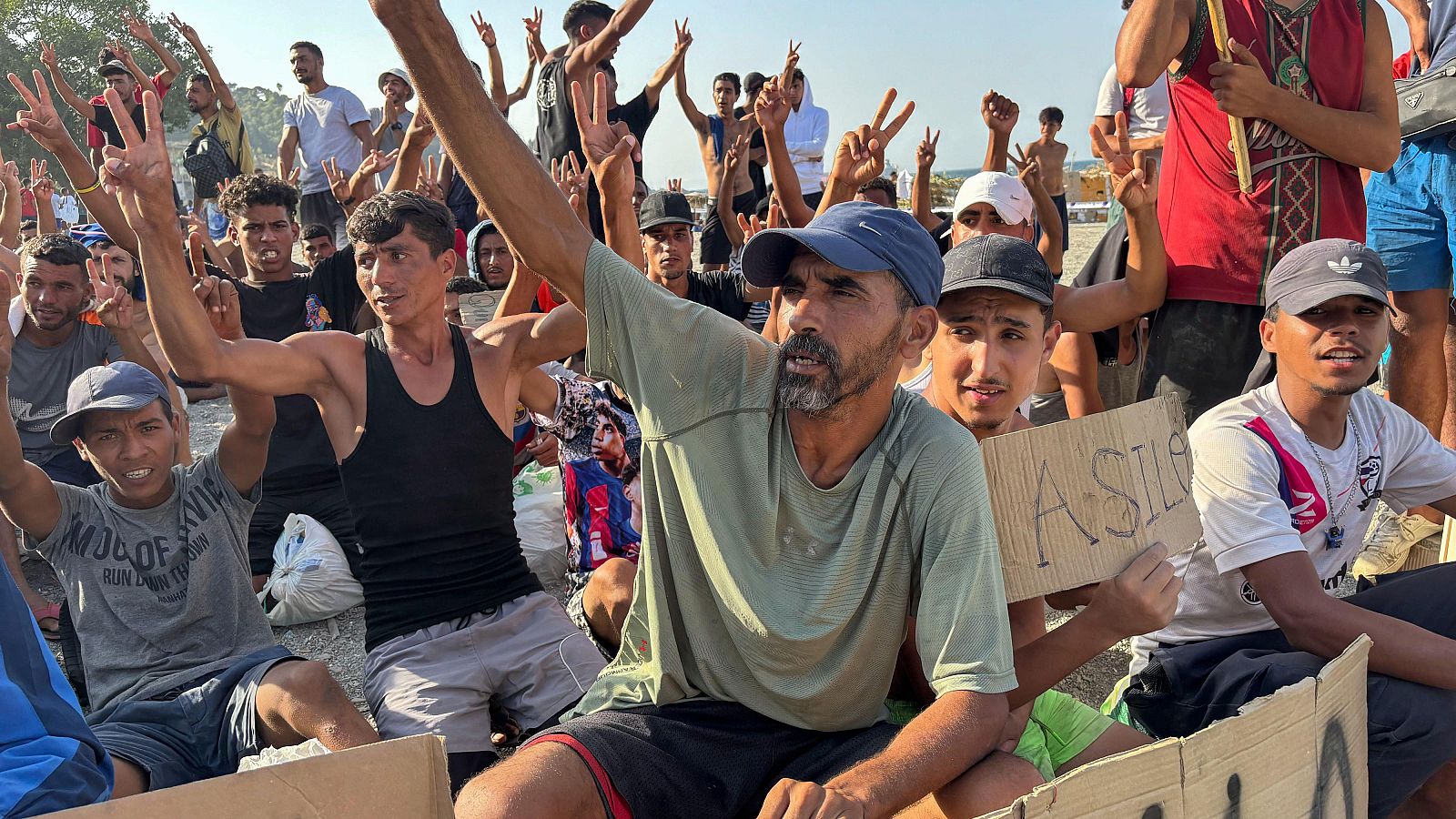 Hombres en terreno polvoriento levantan brazos y hacen el signo de la "V", uno sostiene cartel "ASILE".