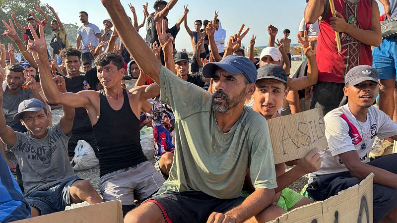 Hombres en terreno polvoriento levantan brazos y hacen el signo de la "V", uno sostiene cartel "ASILE".
