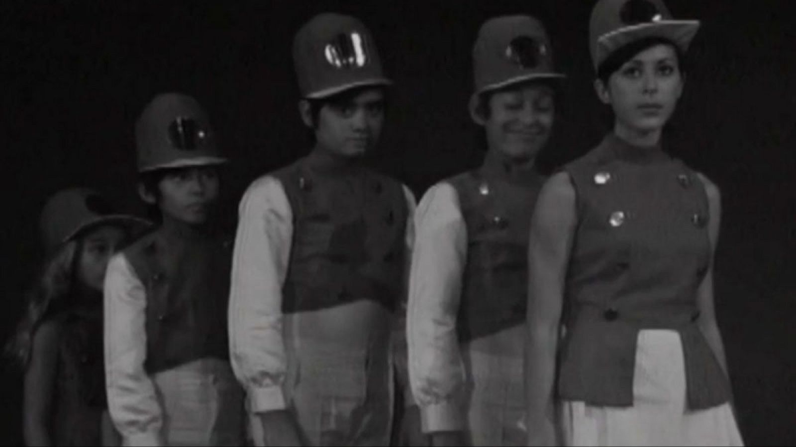 Cinco jóvenes con uniformes y gorras posan en fila. Imagen en blanco y negro de aspecto antiguo, con detalles de chalecos y gorras.