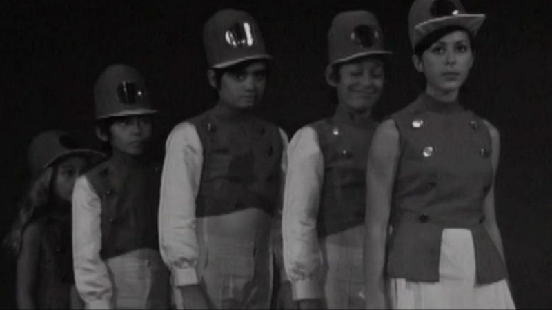 Cinco jóvenes con uniformes y gorras posan en fila. Imagen en blanco y negro de aspecto antiguo, con detalles de chalecos y gorras.