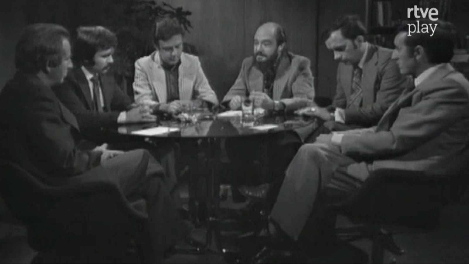 Seis hombres en un estudio de TV, alrededor de una mesa redonda, debatiendo. Presenta a Fernando Jiménez del Oso, conductor de "Más allá". Logo "rtve play" visible. Fotografía en blanco y negro, 1976-1981.