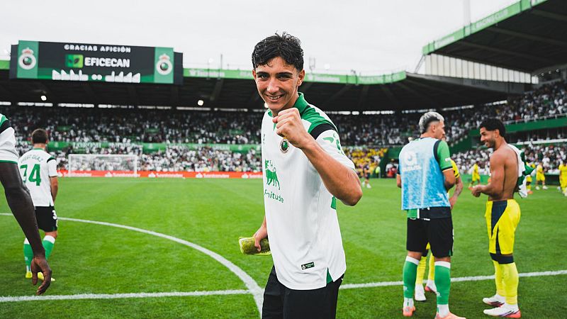Sergio Martínez, canterano, sonríe a cámara con el puño cerrado, vistiendo camiseta blanca con escudo y patrocinador "Frituse". Sostiene una fruta amarilla. Detrás, gradas y pantalla con "GRACIAS AFICIÓN".