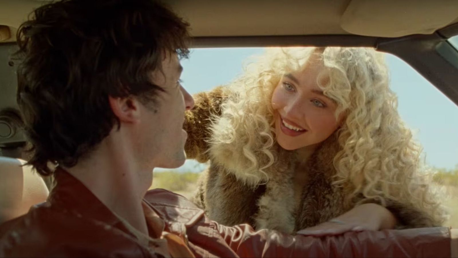 Sabrina Carpenter, con cabello rubio voluminoso y abrigo de piel, sonríe desde la ventanilla de un coche a un hombre joven con chaqueta de cuero. Cielo azul y paisaje árido al fondo.