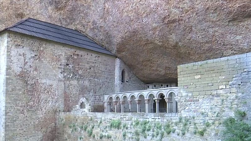 Edificación histórica de piedra, con arcos de medio punto y tejados oscuros, construida bajo un gran saliente de roca natural. Integración arquitectónica y paisajística.
