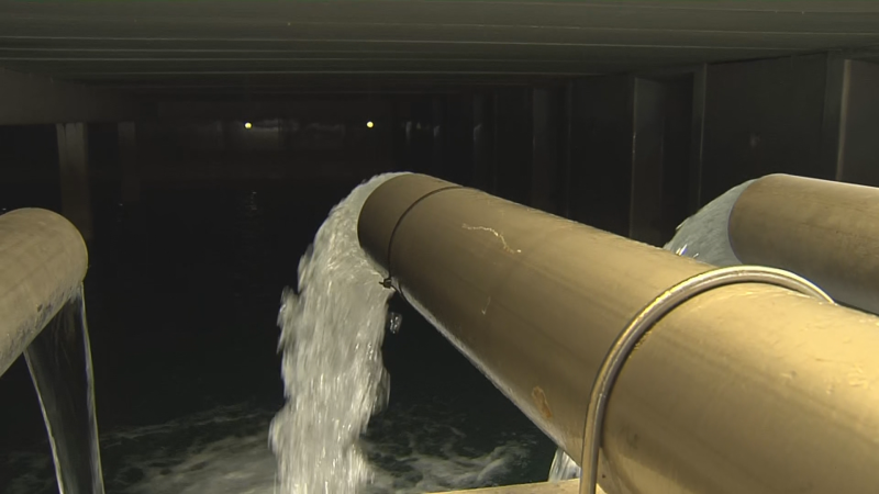 Tuberías de gran diámetro vierten agua con fuerza en un canal oscuro, creando espuma y turbulencia. Infraestructura de hormigón en un ambiente de baja luminosidad.