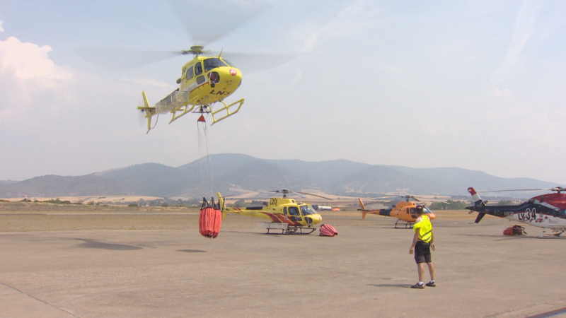 Helicóptero amarillo en vuelo con "Bambi bucket" rojo sobre aeródromo con otros helicópteros y personal de apoyo. Montañas al fondo.