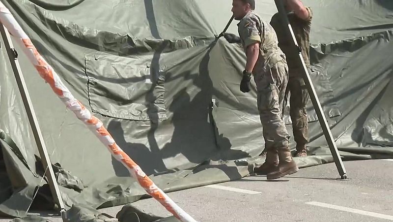 Dos individuos con uniformes de camuflaje montan una tienda verde oliva sobre asfalto. Se aprecian postes metálicos y un poste blanco con marcas naranjas en primer plano.
