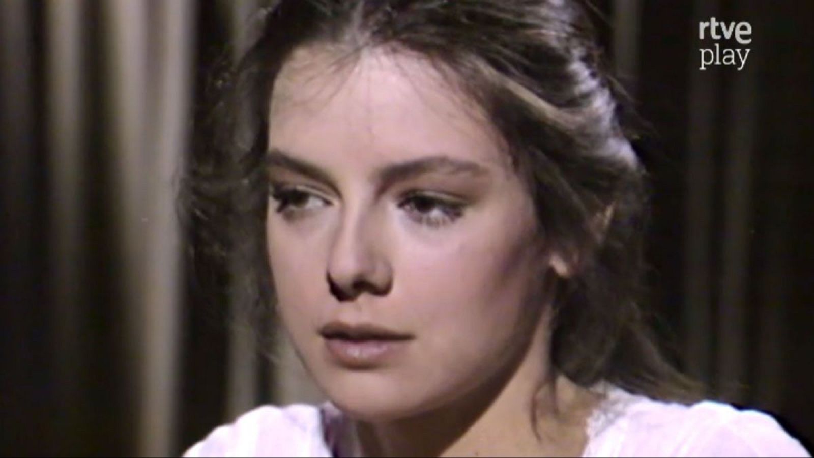 Lola Forner en su juventud, primer plano con expresión serena. Cabello castaño oscuro, vestimenta clara con encaje. Logo "rtve play" visible.