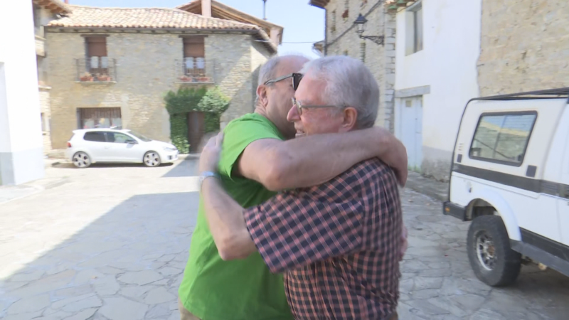 Dos hombres mayores se abrazan en una calle de pueblo. Uno con camiseta verde y gafas de sol, el otro con camisa a cuadros y gafas de vista. Edificios de piedra y coches al fondo.