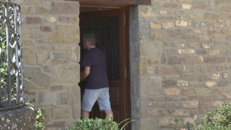 Hombre de espaldas en puerta rústica de piedra, con reja metálica y verja de hierro forjado. Entorno exterior con vegetación.