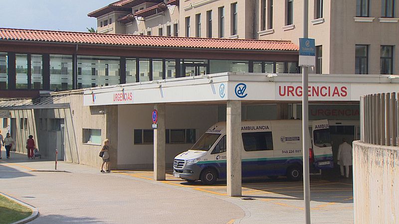 Entrada de Urgencias de un hospital en Cantabria con ambulancias y señalización visible. Se aprecian personas y vegetación en el exterior.