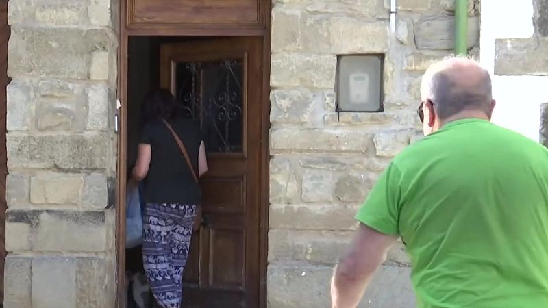 Mujer de espaldas entrando a edificio de piedra, viste camiseta negra y pantalones de elefantes. Hombre calvo con camiseta verde se acerca a la puerta.
