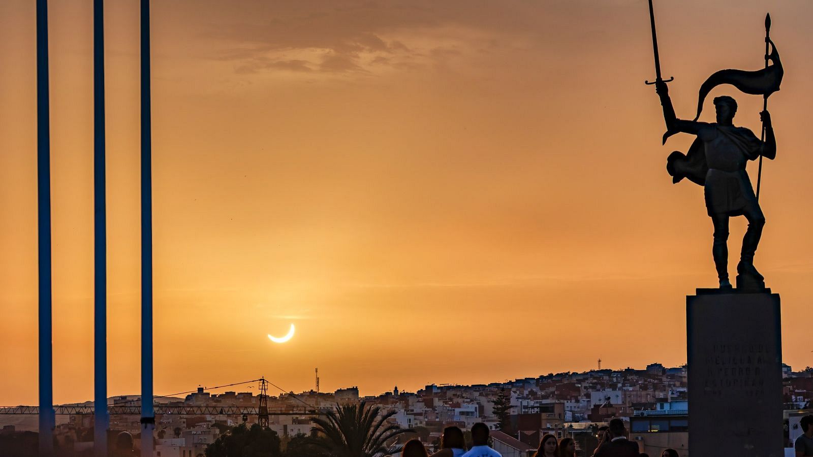 Atardecer con eclipse solar parcial en Melilla. Estatua heroica con espada y estandarte domina el horizonte urbano. Tres postes azules y figuras humanas observan el fenómeno.