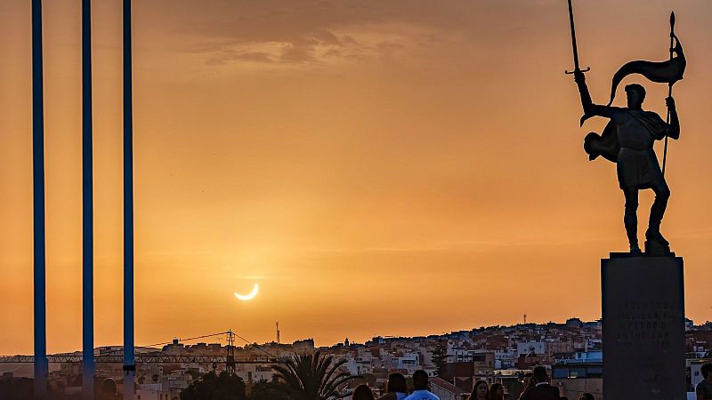 Atardecer con eclipse solar parcial en Melilla. Estatua heroica con espada y estandarte domina el horizonte urbano. Tres postes azules y figuras humanas observan el fenómeno.