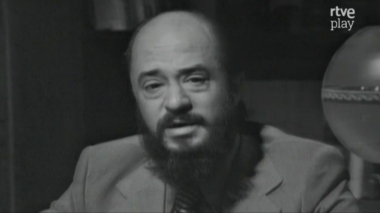 Fernando Jiménez del Oso en primer plano, con barba oscura, traje y corbata. Boca entreabierta, mirada a la izquierda. Fondo oscuro con esfera y logo "rtve play".