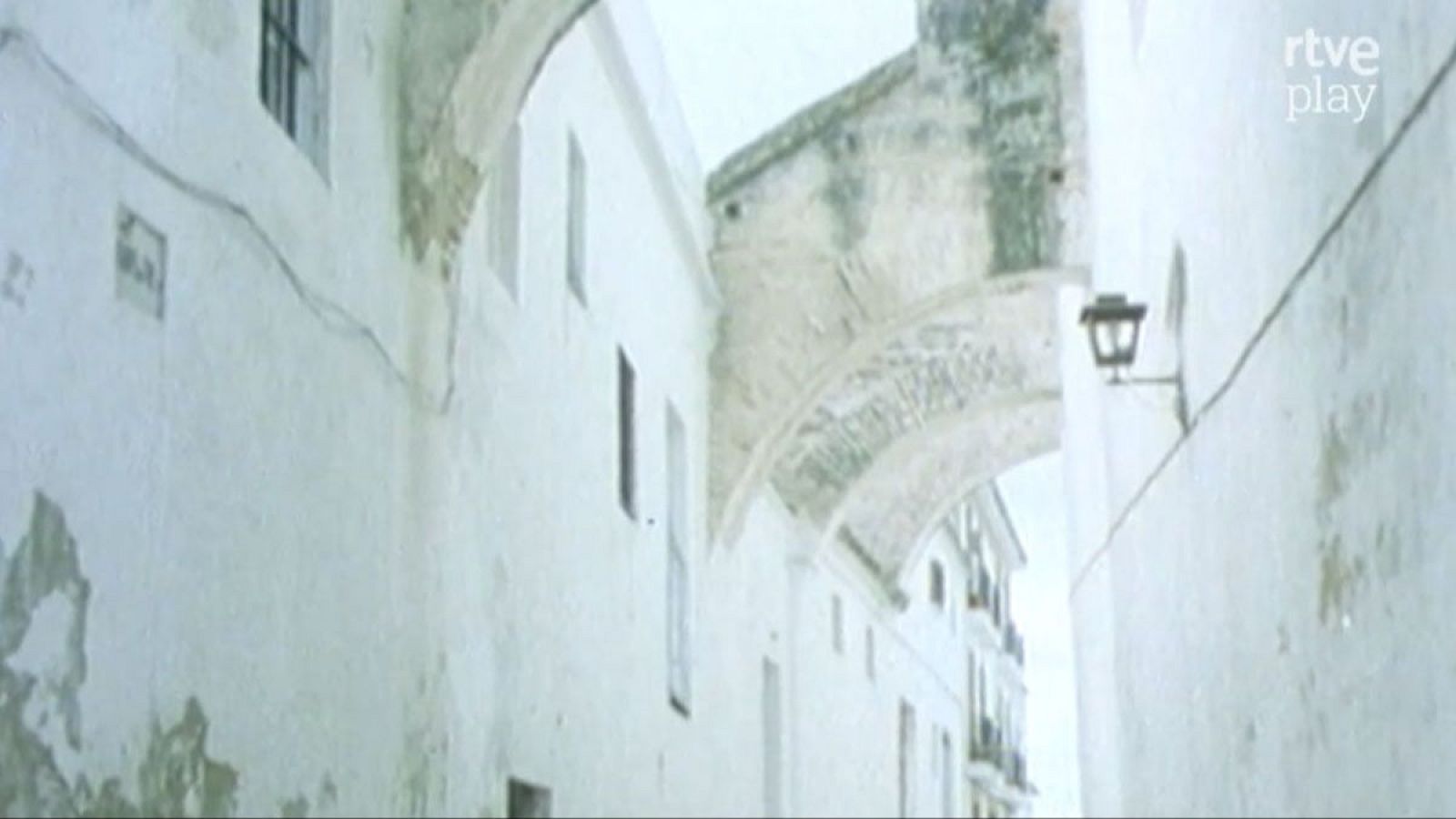 Pasaje andaluz con edificios blancos, arco de mampostería y farola clásica. "rtve play" visible en la esquina superior derecha.