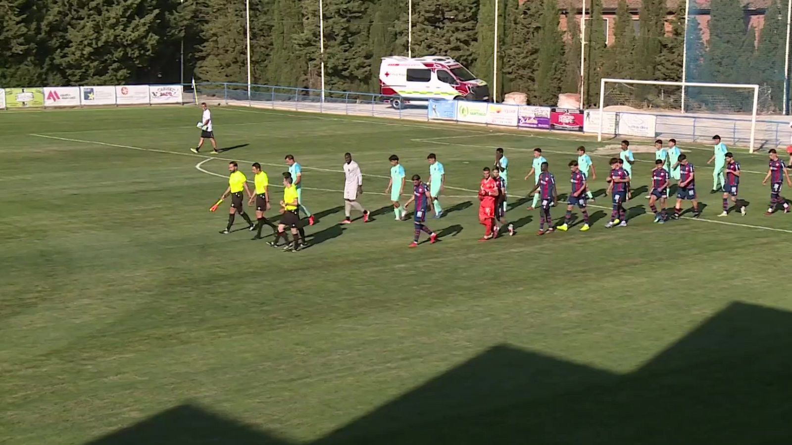 Un partido de fútbol en curso con árbitros y jugadores de dos equipos, uno de burdeos y otro azul claro, en un campo soleado con una ambulancia al fondo.