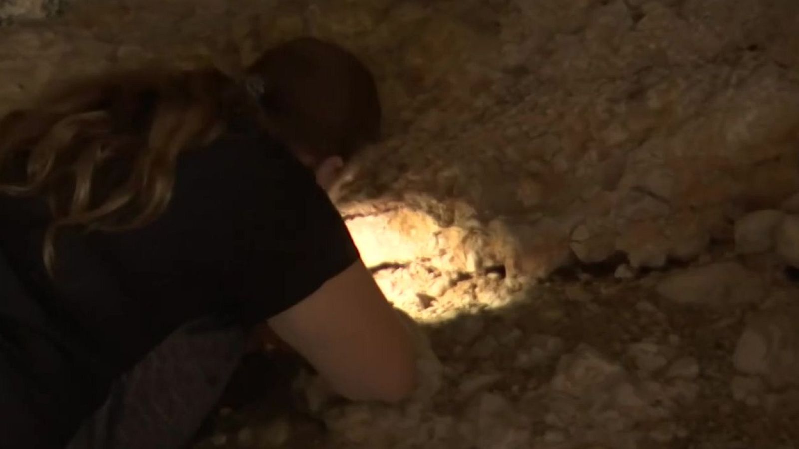 Persona agachada en entorno rocoso oscuro, examinando superficie iluminada por potente haz de luz. Pelo largo castaño recogido en coleta. Atmósfera de exploración.