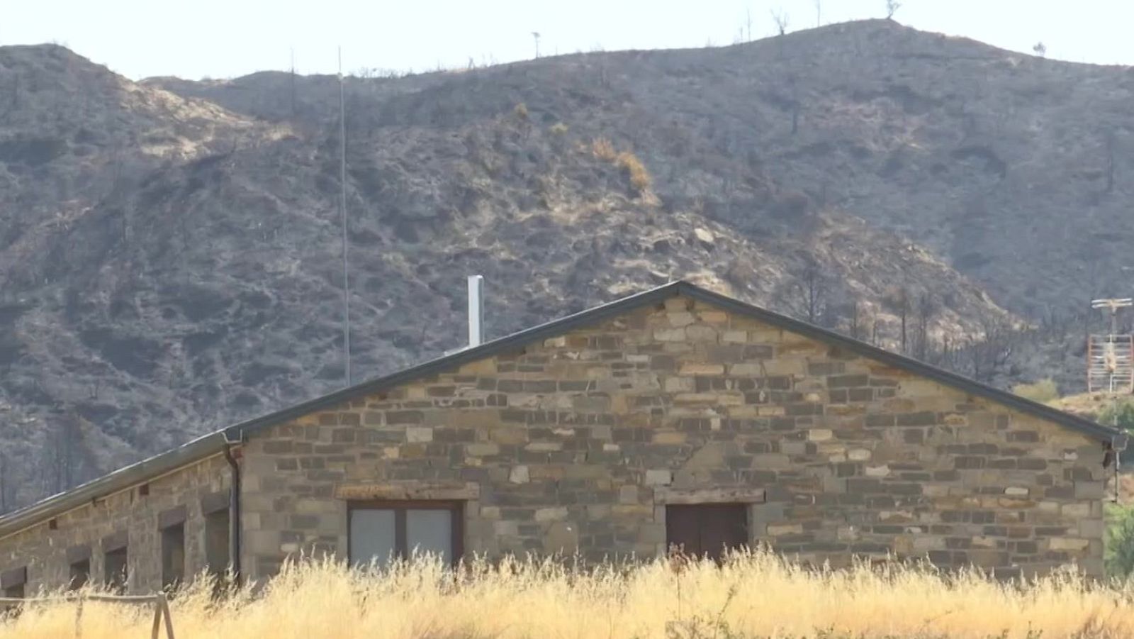 Paisaje rural con edificio de piedra y montaña quemada por incendio forestal. Hierba seca dorada cubre el primer plano.