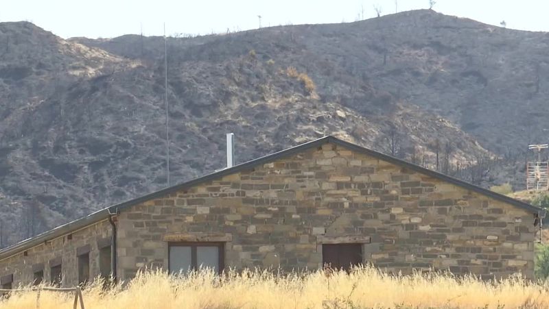 Paisaje rural con edificio de piedra y montaña quemada por incendio forestal. Hierba seca dorada cubre el primer plano.