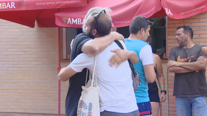 Dos hombres se abrazan bajo un toldo rojo con los textos "CERVECEROS INDEPENDIENTES" y "AMBAR". Otros hombres y una mujer observan en un evento informal.