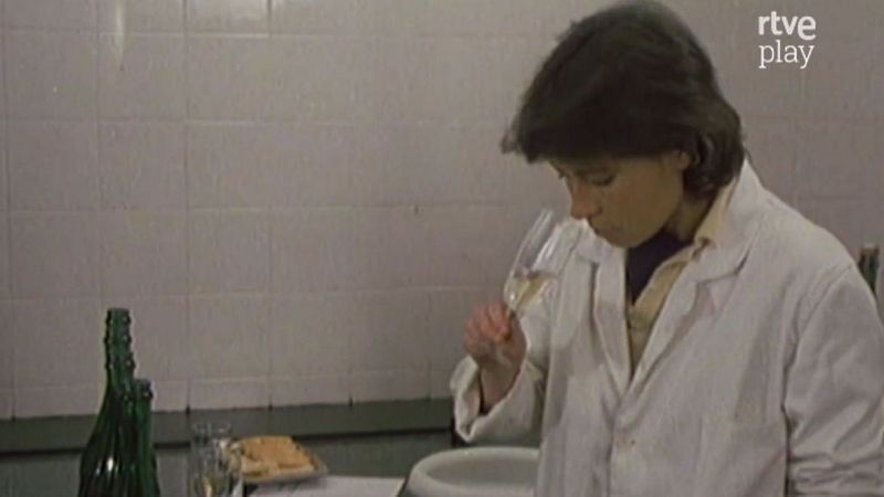 Mujer en bata de laboratorio cata cava, oliendo una copa con l�quido burbujeante. Botella de cava, vaso y aperitivos en mesa. Fondo de azulejos grises. Logo "rtve play" visible.