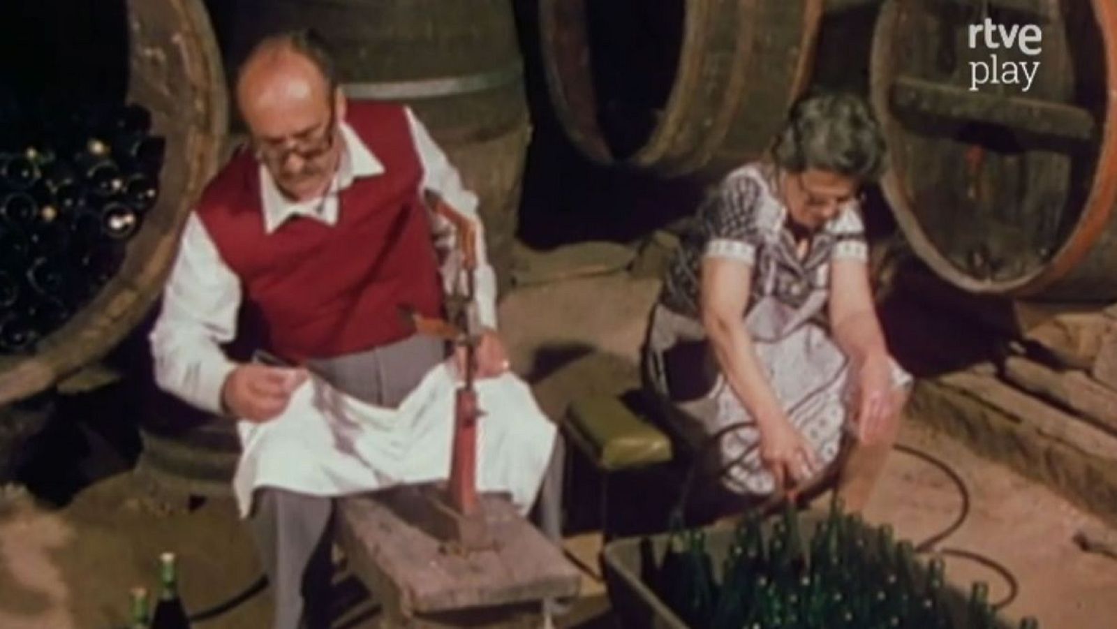 Bodega tradicional con dos personas mayores trabajando en el embotellado. Hombre con máquina roja, mujer organizando botellas verdes. Logo 'rtve play' visible.