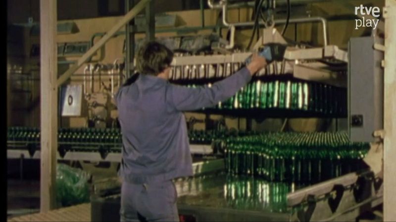 Operario en embotelladora de vino manipula maquinaria con miles de botellas verdes. "rtve play" visible.
