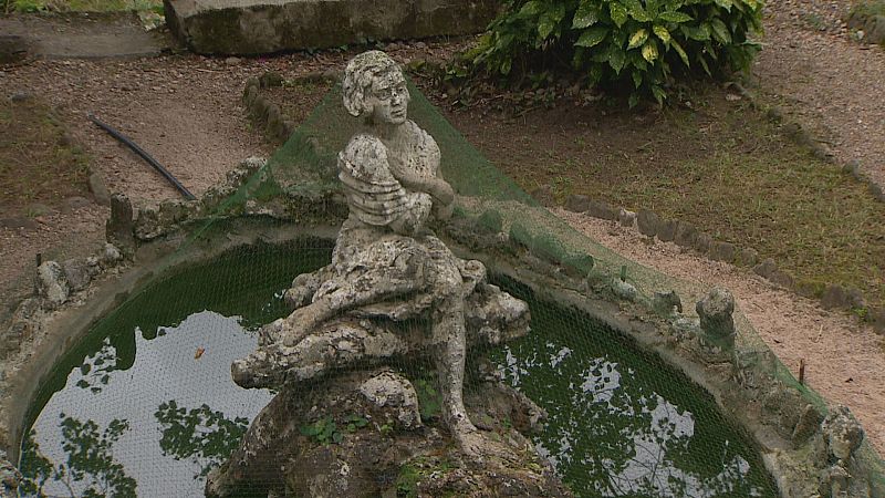 Estatua de piedra erosionada en un estanque verde con red protectora, parte de la Ruta Delibes en Molledo, Cantabria.