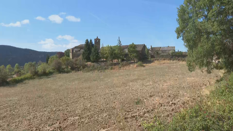 Paisaje rural con campo seco, edificaciones tradicionales y una iglesia con campanario, rodeado de vegetación y montañas bajo un cielo azul.