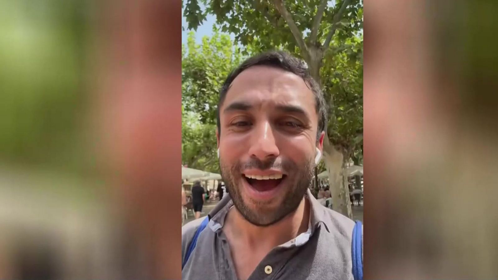 Hombre sonriente con camisa gris, auriculares blancos y correa azul, en un entorno exterior arbolado. Mira a la cámara con alegría.