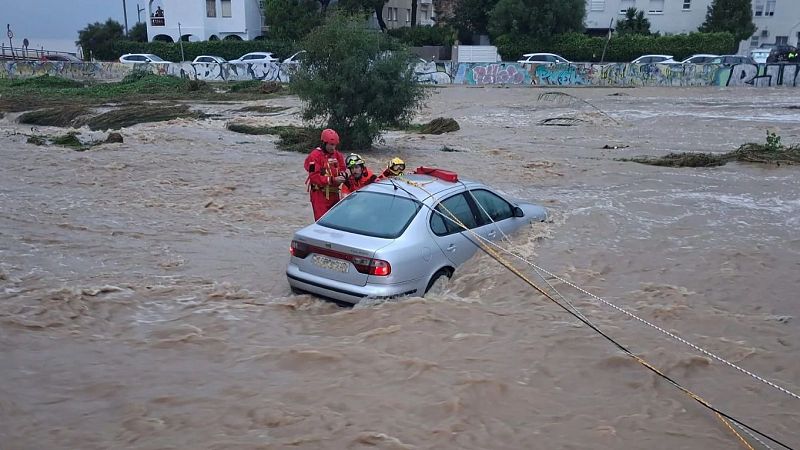 Rescate de un coche plateado en una inundación urbana. Tres rescatistas con uniformes rojos y cascos de colores trabajan en la operación.