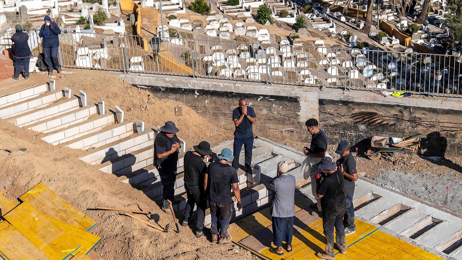 Vista aérea de un cementerio con zona antigua y nueva en construcción. Un grupo de hombres observa una excavación con bloques de hormigón, uno de ellos en actitud reflexiva.
