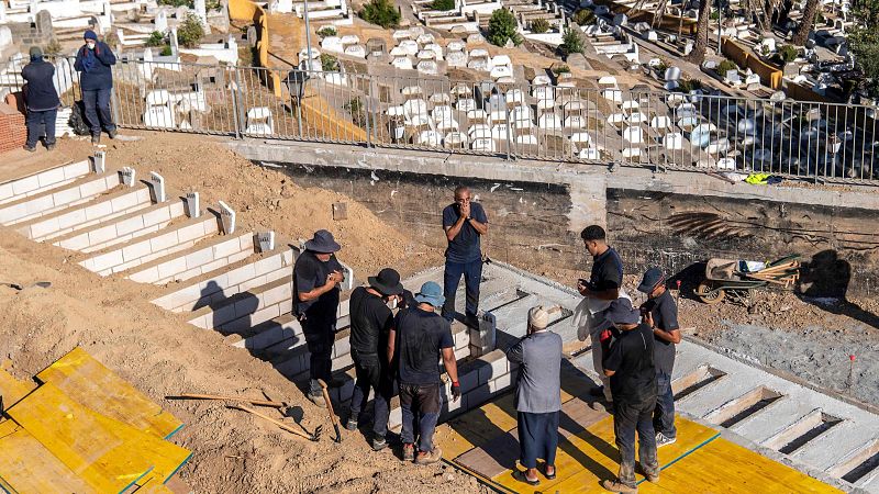 Vista aérea de un cementerio con zona antigua y nueva en construcción. Un grupo de hombres observa una excavación con bloques de hormigón, uno de ellos en actitud reflexiva.