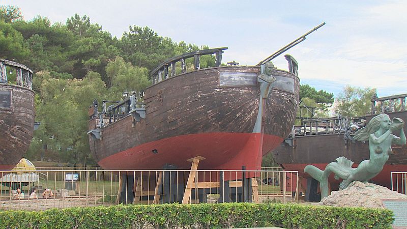 Galeones de madera y una estatua de sirena en exhibición exterior, con un bosque de pinos al fondo y un cielo azul.
